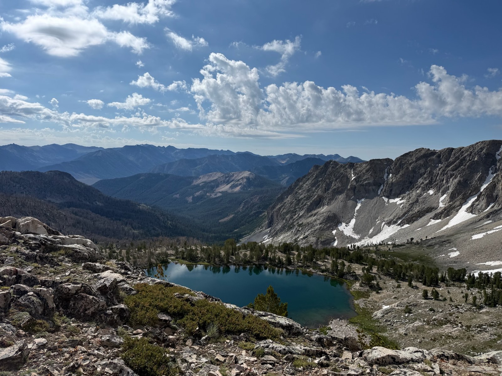 A clear alpine lake sits in a rocky basin in the Idaho backcountry, ringed by evergreens with snow-streaked granite ridges on the right and layered mountains fading into the distance under a bright, cloud-scattered sky.
